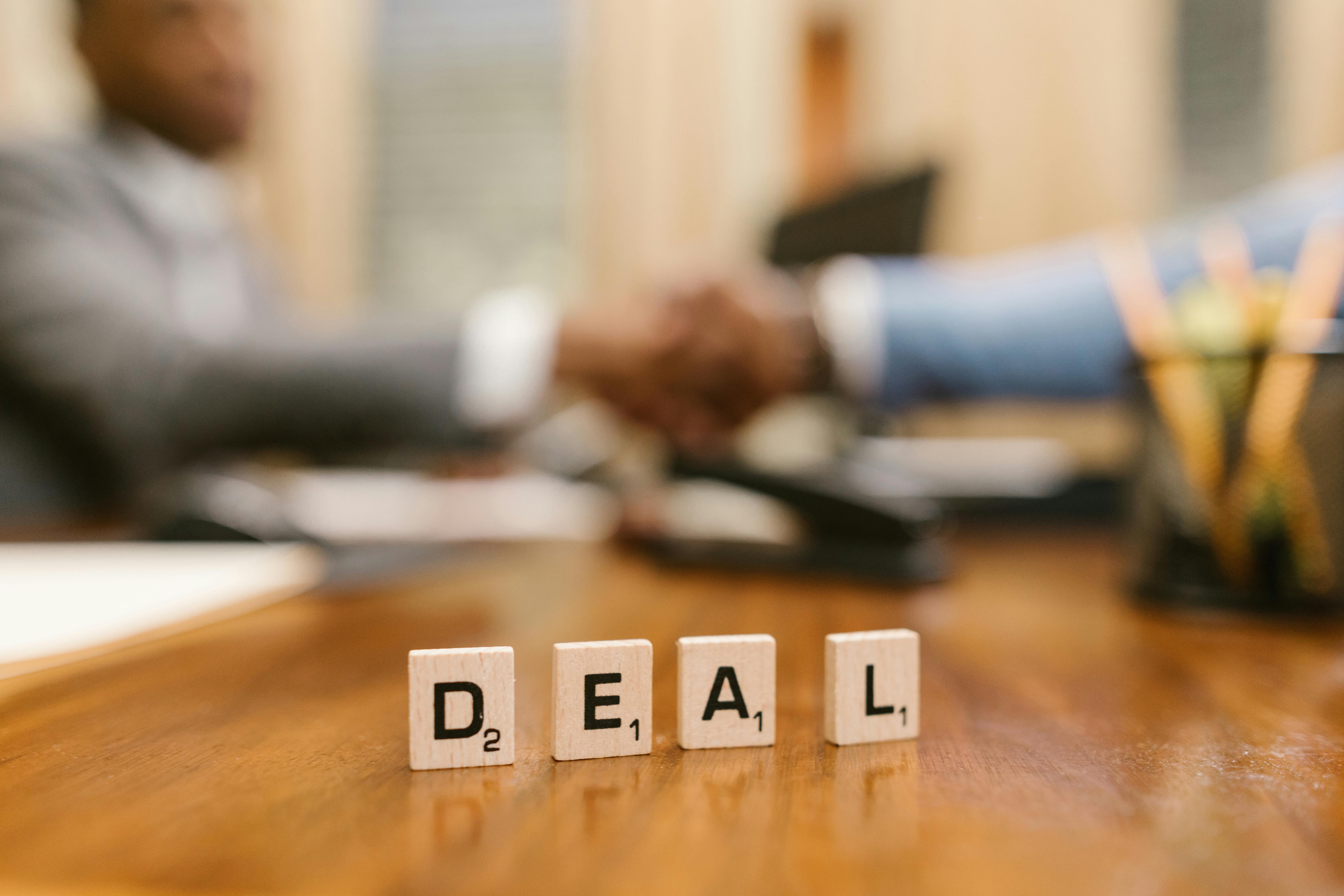 two people shaking hands while the camera's focus is on the word "deal" spelled out in Scrabble tiles