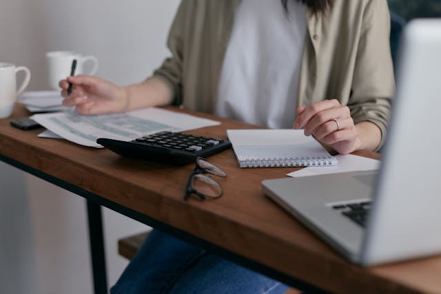 person at wooden desk with forms, a calculator, and a laptop